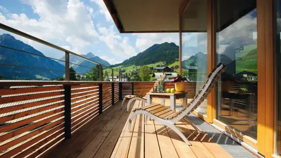 Wooden balcony with chairs and a table, surrounded by mountains and clouds.