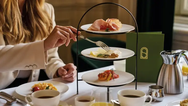 A woman is sitting at a breakfast table, holding a fork in her hand and guiding it to an etagere with cheese, sausage and fish. There is also a menu, a coffee pot, two coffee cups and a glass of orange juice on the table. 