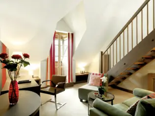 A living area with two armchairs, a small glass table, a desk and a chair. A red vase with roses can be seen in the front left of the picture and a wooden staircase leads upwards in the background.