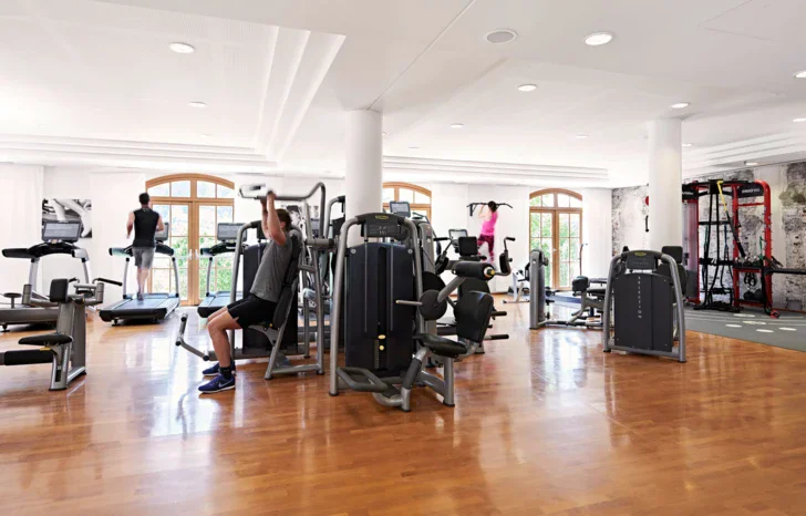 People exercising in a well-equipped gym with various machines, wooden floors and white walls and ceiling. 