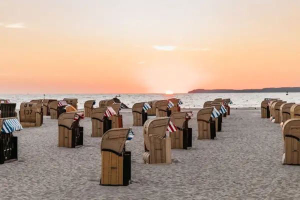 A beach with a gently undulating sea full of red and blue beach chairs as the sun rises and bathes the sky in a warm orange. 