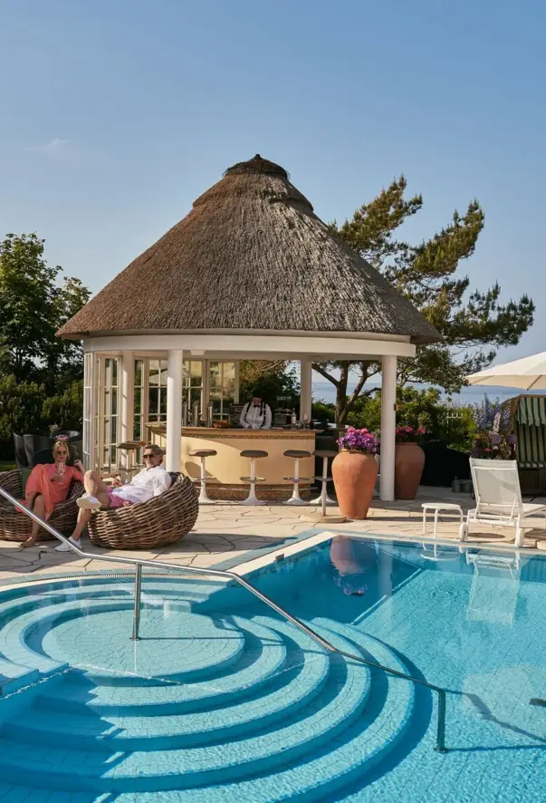 A couple sits in woven armchairs next to an outdoor pool near a bar with a thatched roof. An employee is working at the bar, which has a glass front on one side and bar stools around it. 