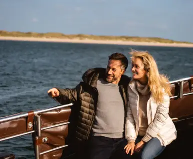 A couple sits arm in arm, smiling on an open boat. The man has his finger pointed at something outside the picture. In the background are dunes and the sea. 
