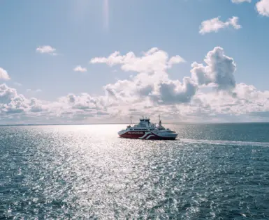 A ferry sails on the open sea over deep blue water. The warm sun is reflected in the sea, the sky is partly covered by light clouds. 