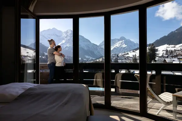 A couple are standing in front of a large window overlooking a snow-covered mountain landscape.
