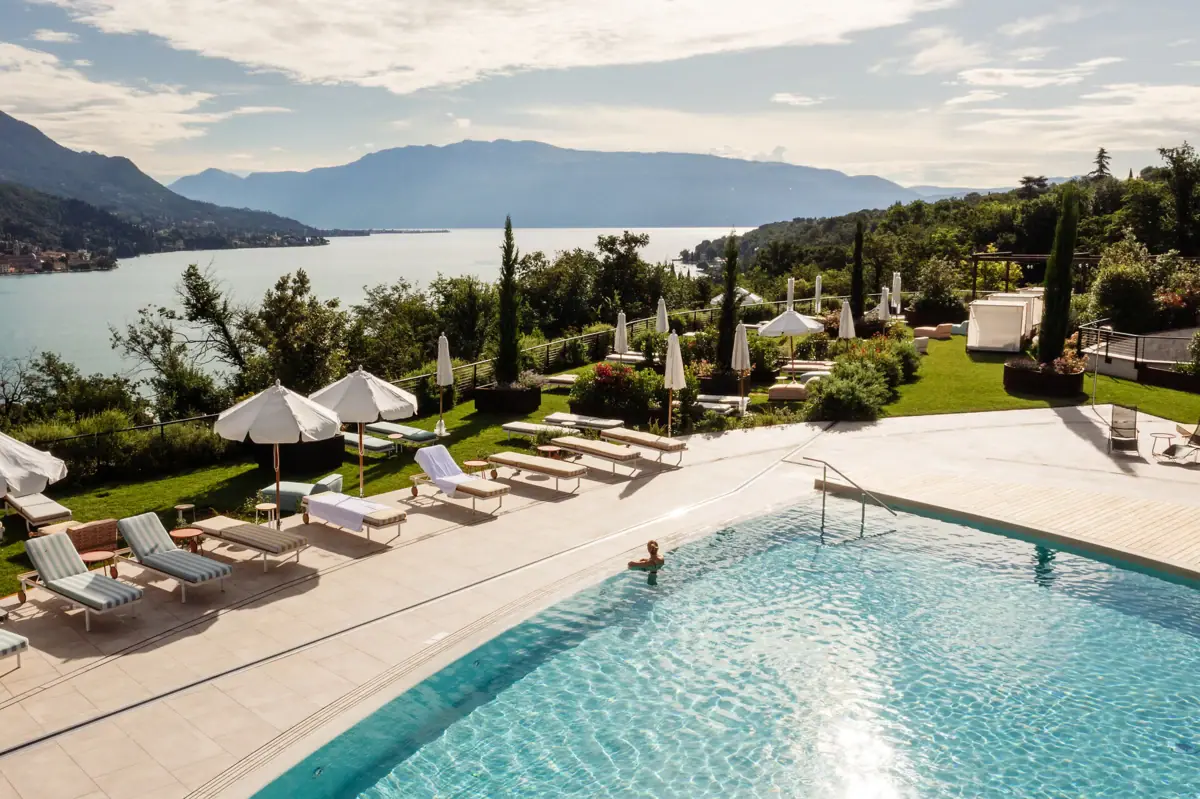 Swimming pool at the A-ROSA on Lake Garda An outdoor swimming pool with crystal-clear water, surrounded by trees, with a mountain and a lake in the background.