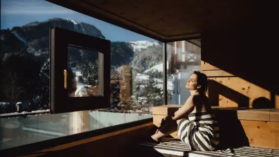 A woman sitting in a wooden sauna with a big window front overlooking snowy trees and mountains. 
