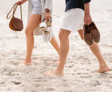 A man and a woman walk barefoot on the beach in a summer outfit.