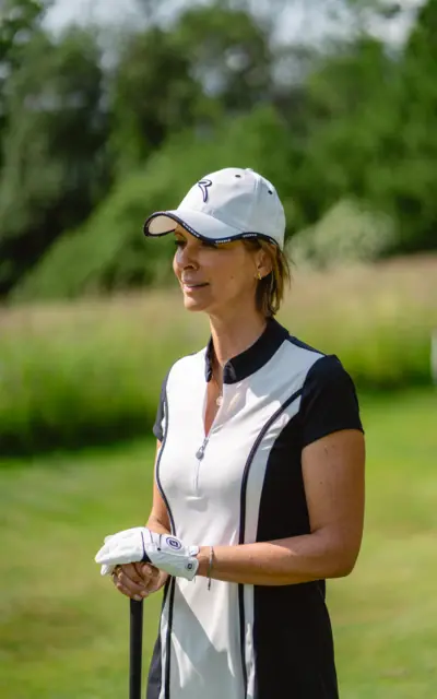 Woman with white cap and white shirt standing on a meadow playing golf.