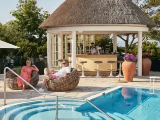 A man and a woman sit in wicker chairs next to a swimming pool and a pool bar.