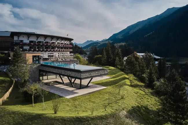 A-ROSA Ifen Hotel Kleinwalsertal A building with a swimming pool in the middle, surrounded by mountains and blue skies.