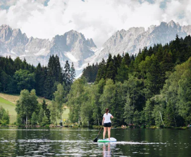 A woman is standing on a stand up paddling board on a quit lake. Trees and mountains are rising in the back. 