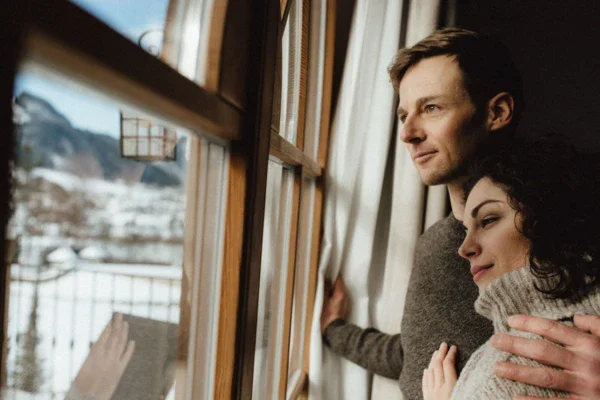 A couple standing arm in arm infant of a window looking out onto the snowy landscapes. 