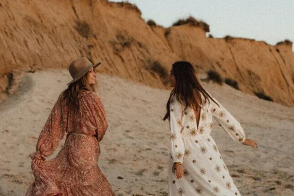 Two women in long summer dresses walk happily along a beach bathed in warm light by the setting sun. 