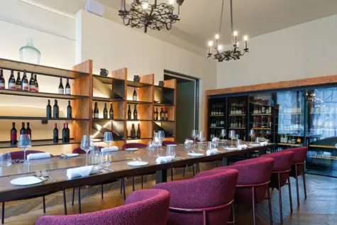 A room with wine bottles and glasses on a table, surrounded by chairs and with shelves in the background.