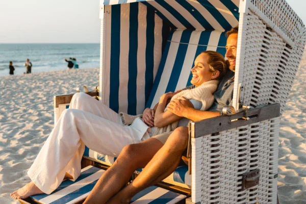 A man and a woman sit smiling, arm in arm, in a white and blue striped beach chair at sunset. The sun shines warm and golden on their skin.