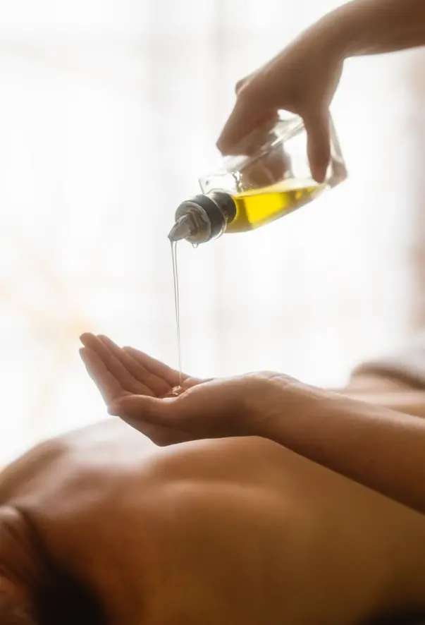 In the background, a woman lies on a massage table and in front of her a hand pours oil from a small glass bottle into another hand.