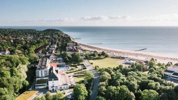 An aerial photo showing the coastal town of Travemünde in bright sunshine. A large white building can be seen in the foreground surrounded by well-kept green areas. A dense row of trees separates the building from a paved road that runs through the middle of the picture. Beyond this, the beach stretches along the coast with orderly rows of sun loungers and parasols.