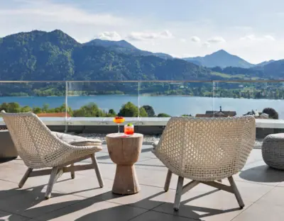 A-ROSA Tegernsee A table and chairs on a patio overlooking a body of water with mountains and clouds in the background.