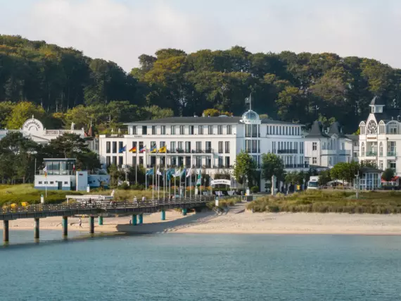 A white building with black-framed windows, a glass dome and a flag on the roof stands in front of a dense beech forest on the coast of Binz. The Baltic Sea can be seen in the foreground as well as a section of Binz beach and the Binz pier, which juts out into the Baltic Sea.