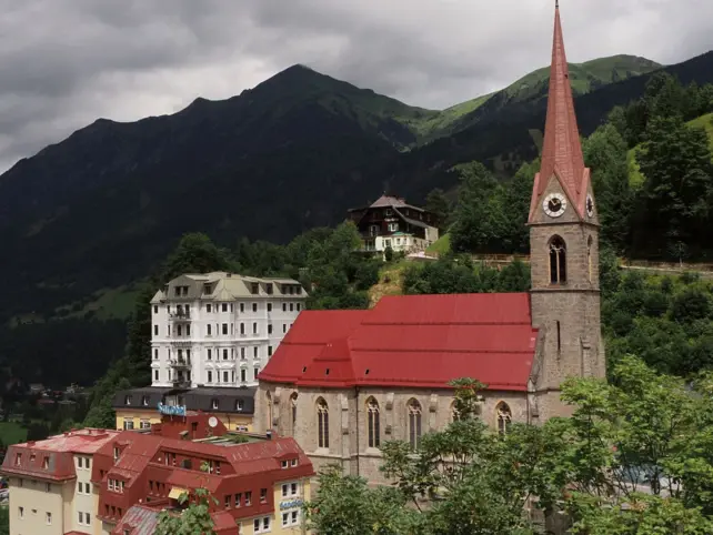 A church on a hill, surrounded by mountains and trees under a cloudy sky.