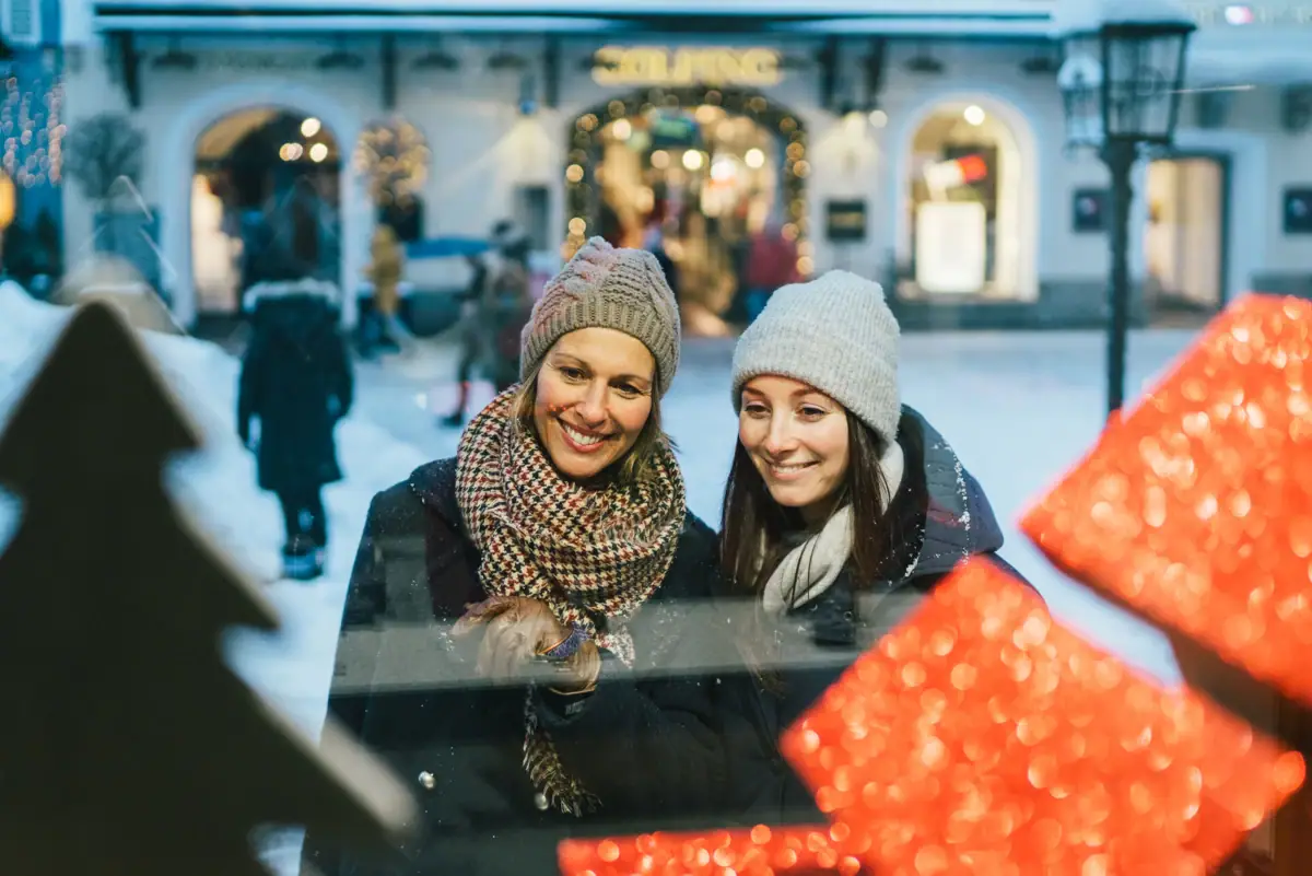 Two women smiling while looking through a window, wearing winter clothing and scarves.