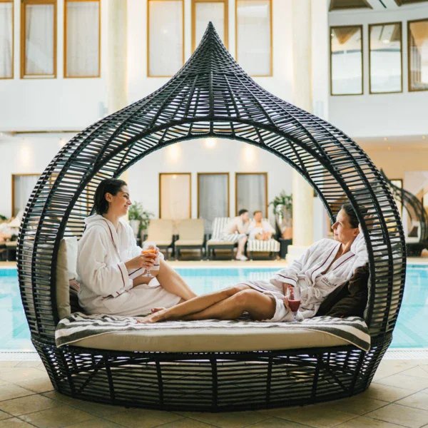 Two woman with drinks and in bathrobes dressed are chatting in a comfortable chair by the pool.