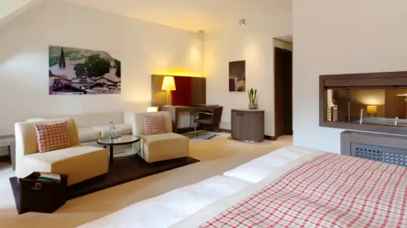 A hotel room photographed from behind the bed. The view falls on a light-colored sitting area with checkered cushions and a desk with a dark chair. 