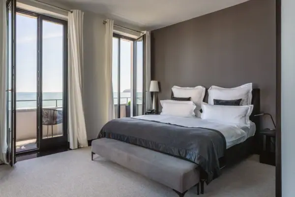 A bedroom with a large window overlooking the ocean, featuring a neatly made bed with pillows and a duvet.