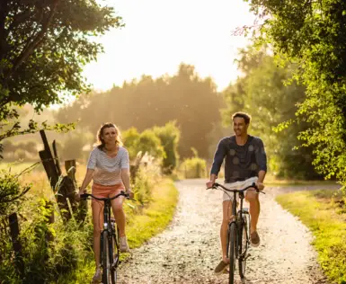 A man and a woman are cycling along a path surrounded by trees and grass.