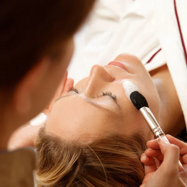 The face of a woman on which a beautician applies a white mask with a brush. 