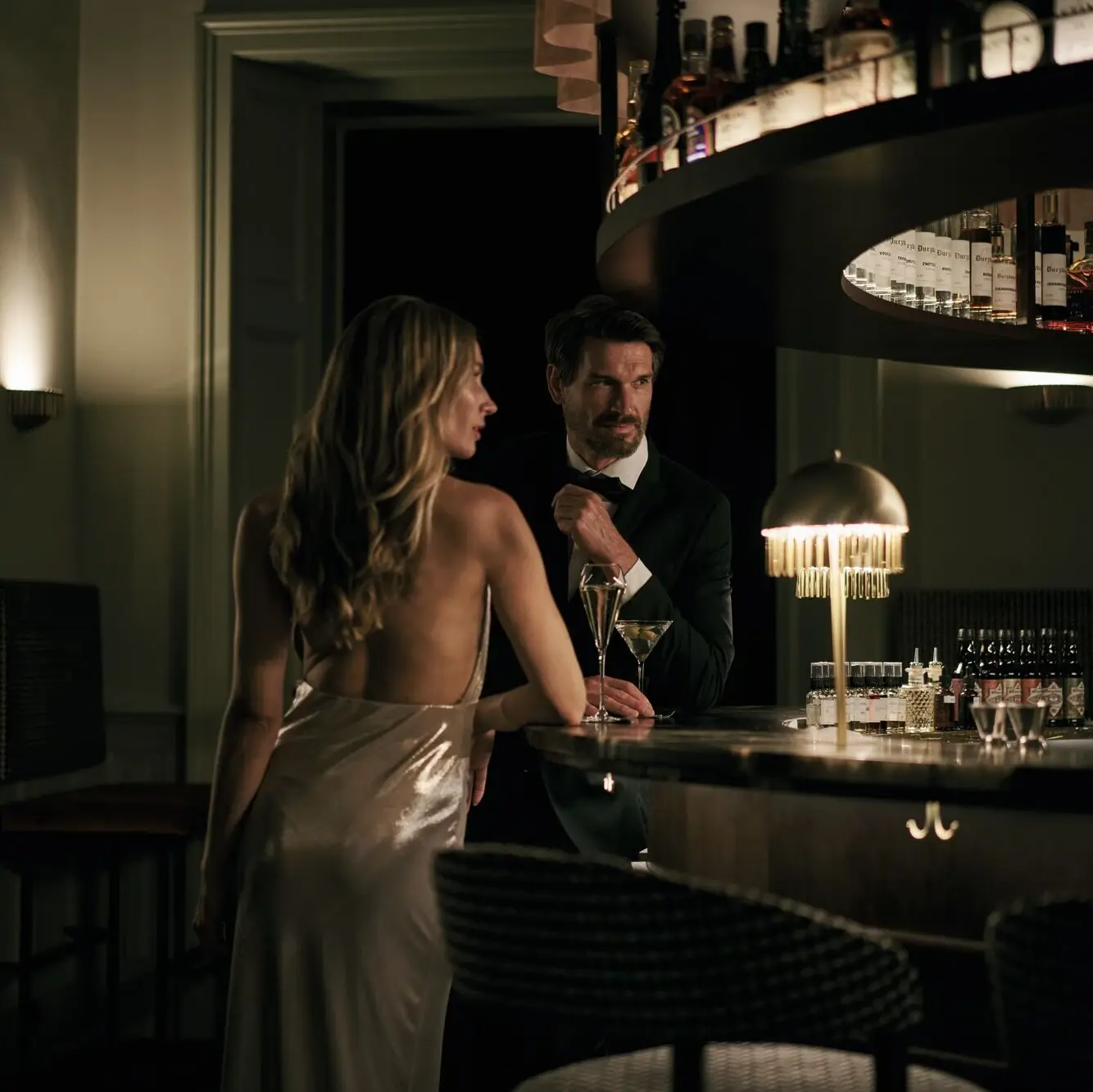 A man and woman standing at a bar indoors, with the woman wearing a dress.