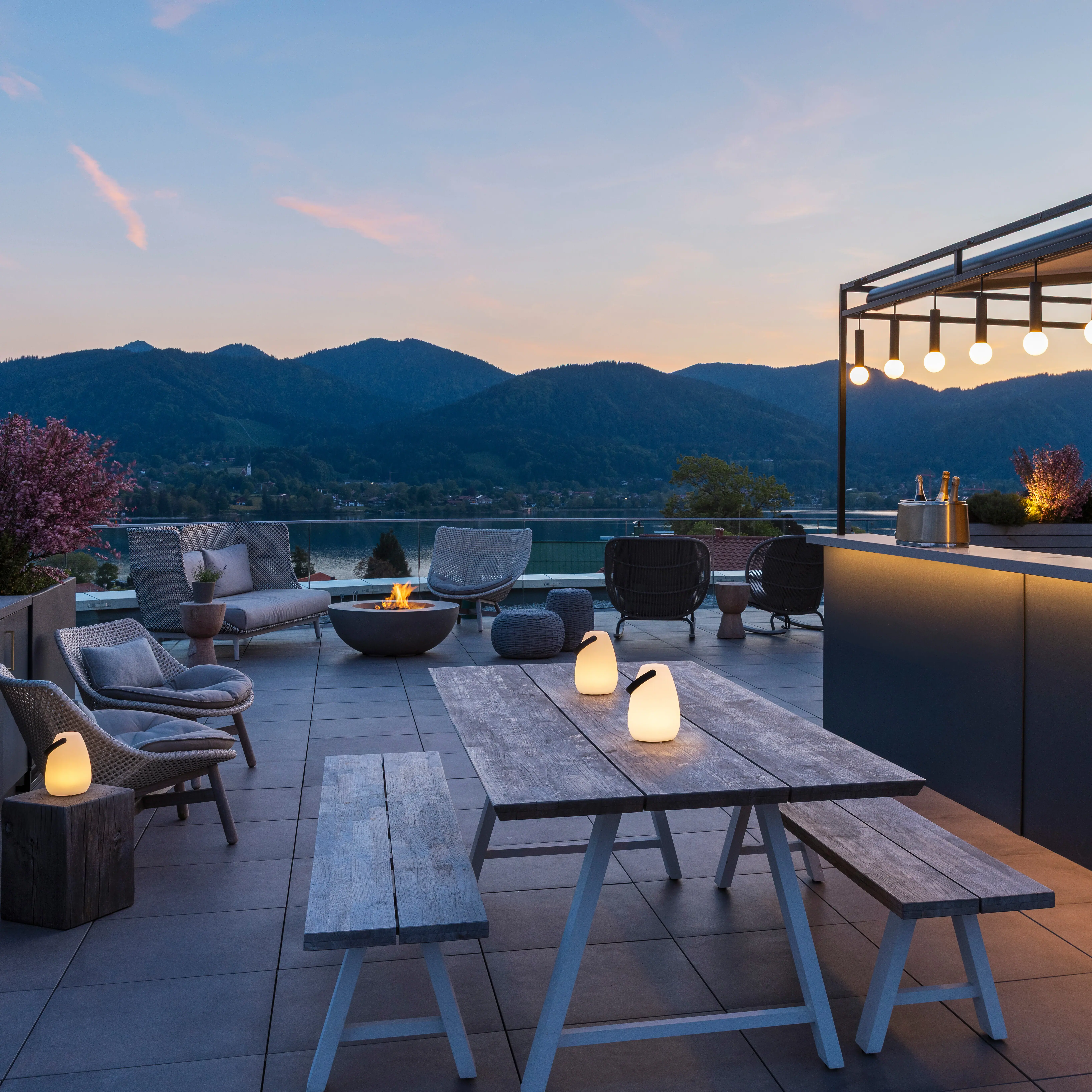 A-ROSA Tegernsee Last Minute A terrace with a table and chairs, a bar in the background and a view of a lake.