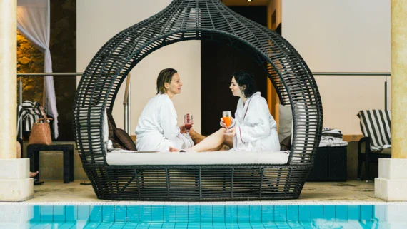 Two women with drinks and dressed in bathrobes are chatting in a comfortable seat by a pool. 