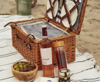 Picnic basket with wine and fruit on a blanket outside.