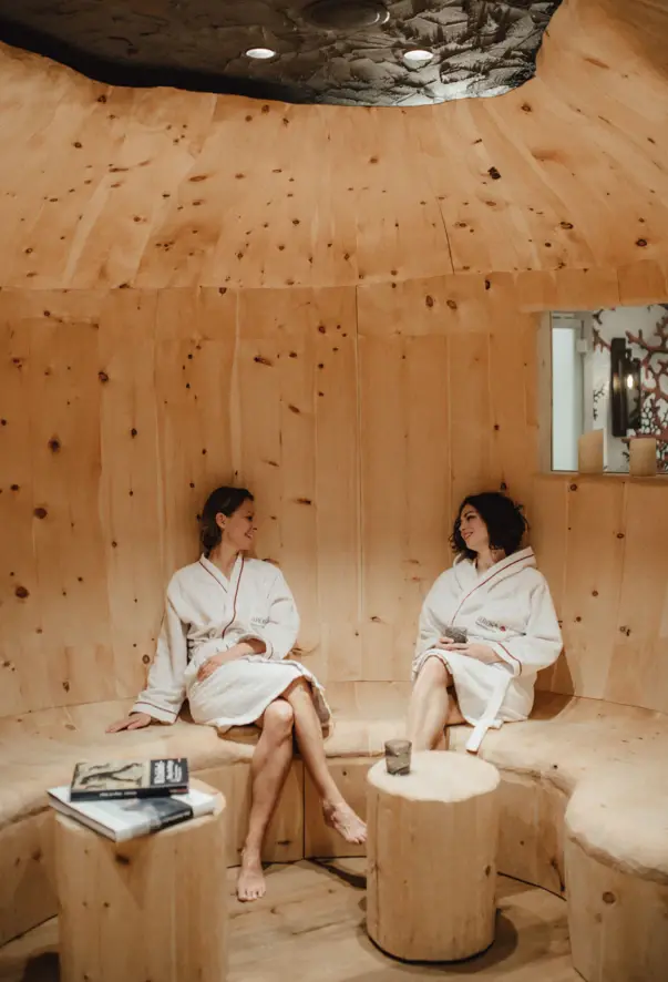 Two woman in robes are sitting in a full wooden room. Little tables are placed infront of the seating both woman are set on and the light is warm. 