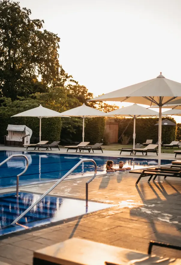 A couple swim in an elegant outdoor pool, surrounded by sun loungers and umbrellas as the sun goes down.