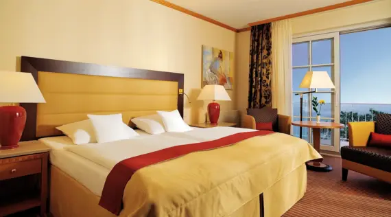 Hotel room with large bed, bedside tables and stylish lighting with a view of the sea through the window.