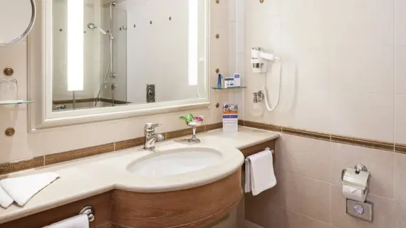 A bright and inviting bathroom with a large mirror above a wooden washbasin. The washbasin has a marble top and a chrome fitting. There is modern lighting on the wall and various bathroom utensils and fresh towels are available. A transparent shower cubicle with a white shower curtain is visible in the background. The neutral tones and clean lines give the room a sense of order and cleanliness.