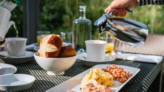 A person pouring coffee into a mug on an outdoor breakfast table with food.