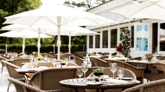 Elegantly laid tables on an outdoor terrace with white parasols and green trees in the background. 