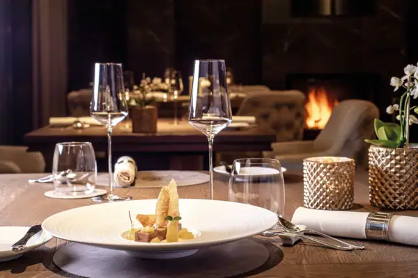 Ein Tisch mit einem exquisiten Gericht auf einem Teller und Weingläsern in einem Innenraum vor einem Kaminfeuer im Hintergrund..