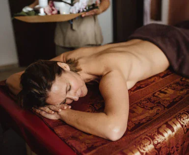 A woman is laying on massage table with a masseuse in the back carrying a board with different products. 