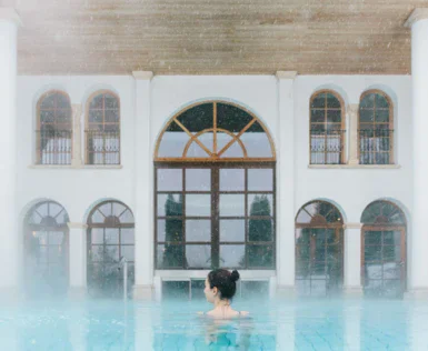 A woman is swimming in a warm steaming outdoor pool facing the entrance to the indoor area with multiple big windows. 