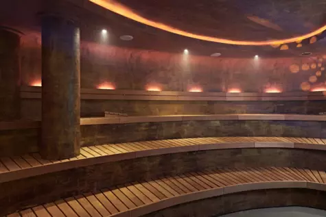 A sauna with wooden seating and stone details on the walls, ceiling and columns. Small lights fill the room with warm light and steam is hanging in the air. 