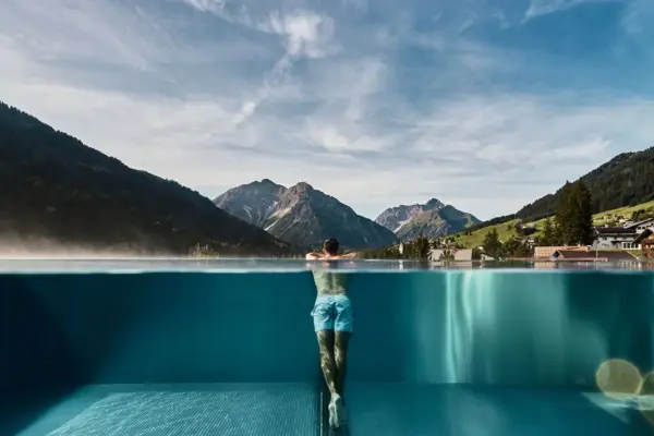 Man leaning on the edge of an infinity pool with a view of a place surrounded by a green mountain landscape in the background.