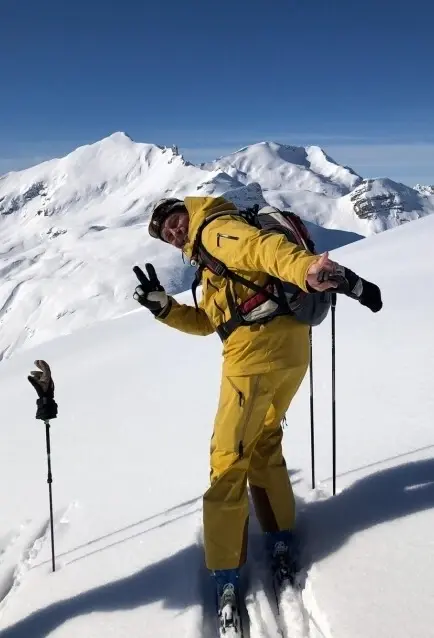 Man in yellow ski suit standing on a snowy mountain with ski equipment and holds the peace sign up.