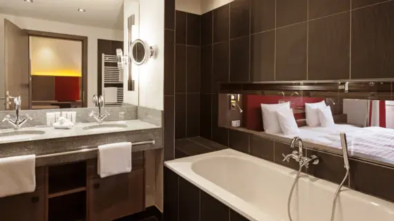 A bathroom with dark tiles, a bathtub and a washstand with two washbasins. A bed in the next room can be seen through a narrow window above the bathtub.