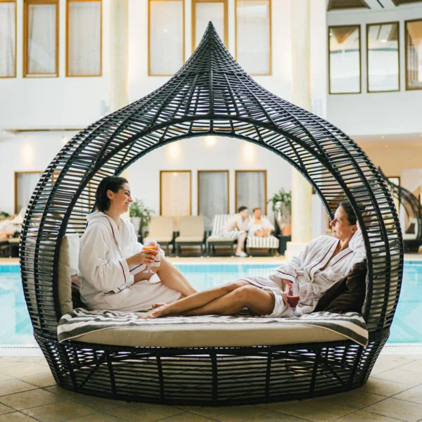 Two woman in robes and with a drink in their hands are sitting in a lounge chair by a pool.