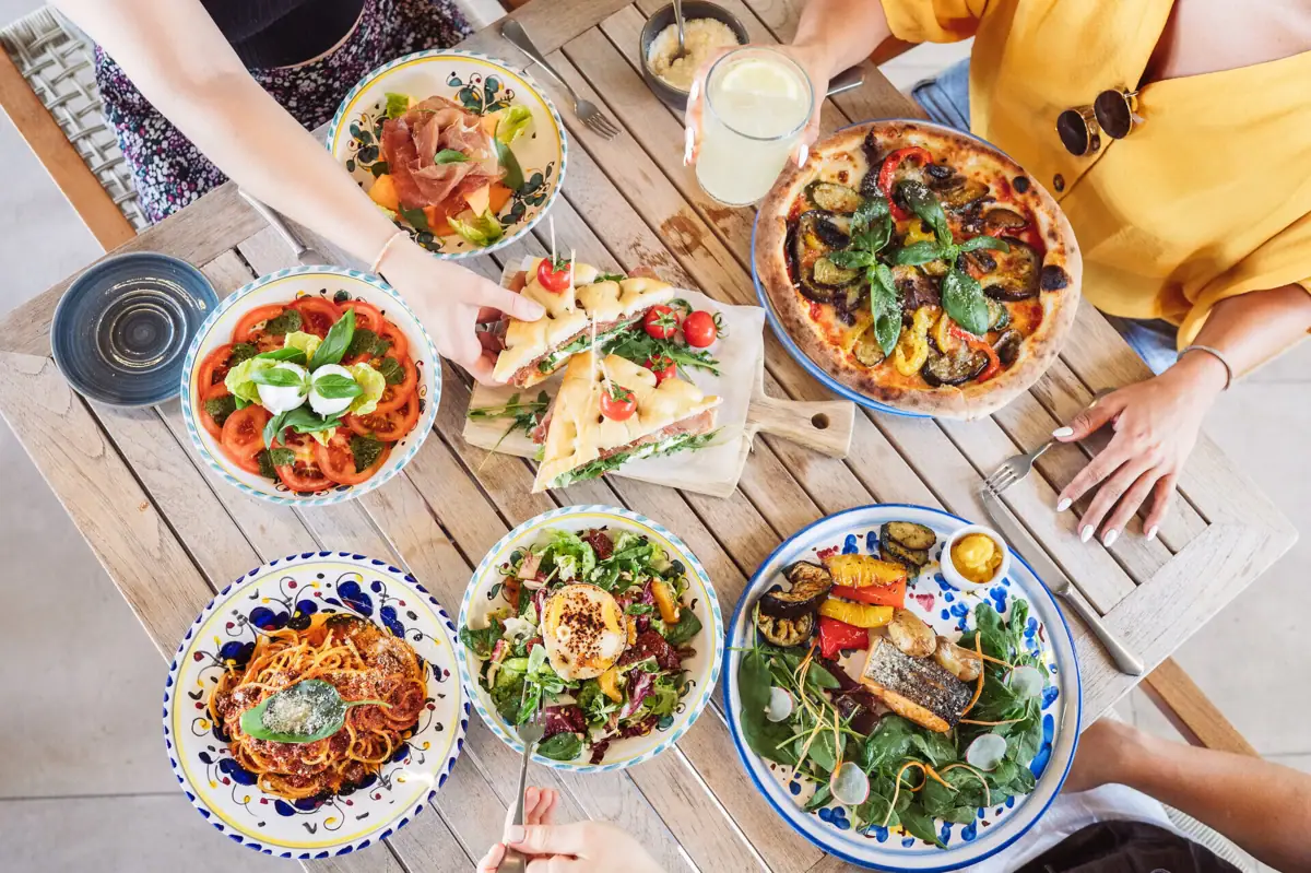 Eine Gruppe von Menschen isst gemeinsam an einem Tisch. Es gibt Pasta, Tomate Mozzarella, vegetairsche Pizza, Lachs mit Salat. Eine Frau nimmt sich gerade ein Focaccia.